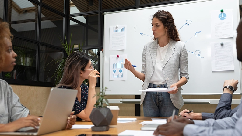 Concentrated multiracial millennial employees listening to confident caucasian team leader explaining project details. Focused diverse teammates discussing marketing strategy with mentor at office.