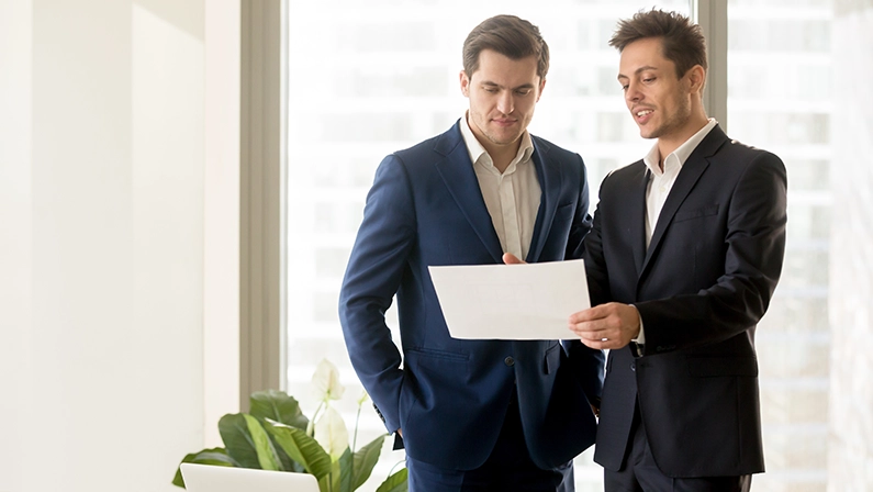 Two smiling businessmen talking about eligible investment, manager presenting financial report showing good work results to satisfied boss, pointing on paper, offering deal, discussing new project