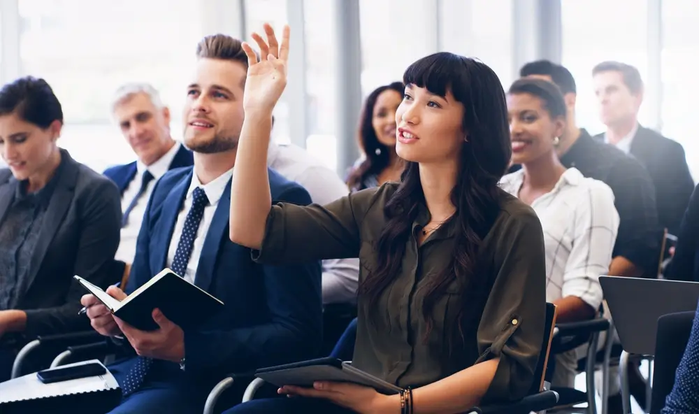 A diverse group of professionals in business attire attending a job training seminar, with one individual raising their hand to ask a question.