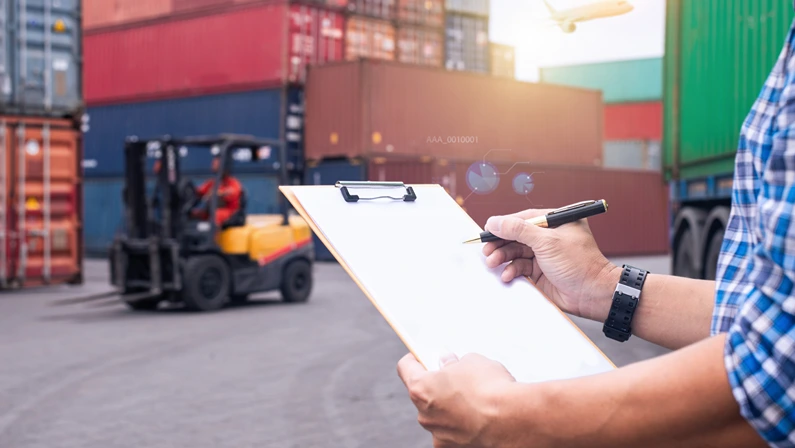 Worker holding a clipboard near shipping containers and forklift, illustrating supply chain risk and logistics management.