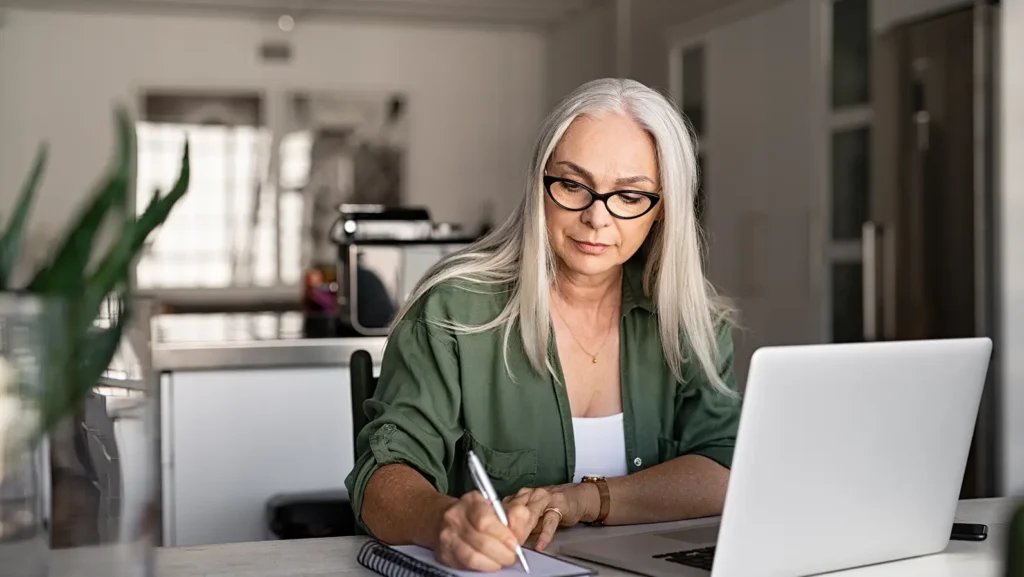 An older woman in glasses taking notes while working on a laptop, exploring the benefits of hiring a grant writer.