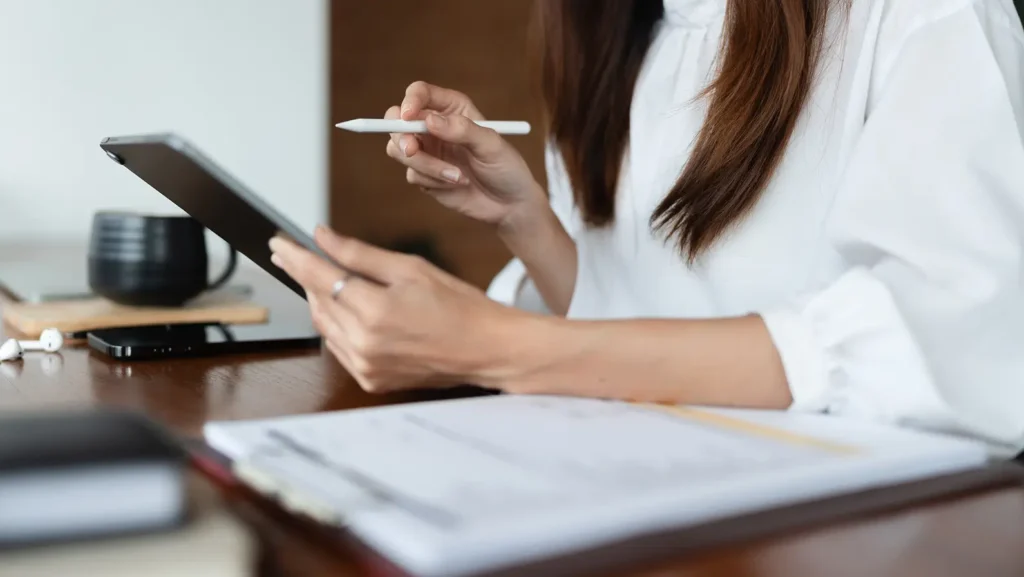 A woman in a white blouse using a tablet and stylus at a desk, researching hiring a grant writer for her organization.