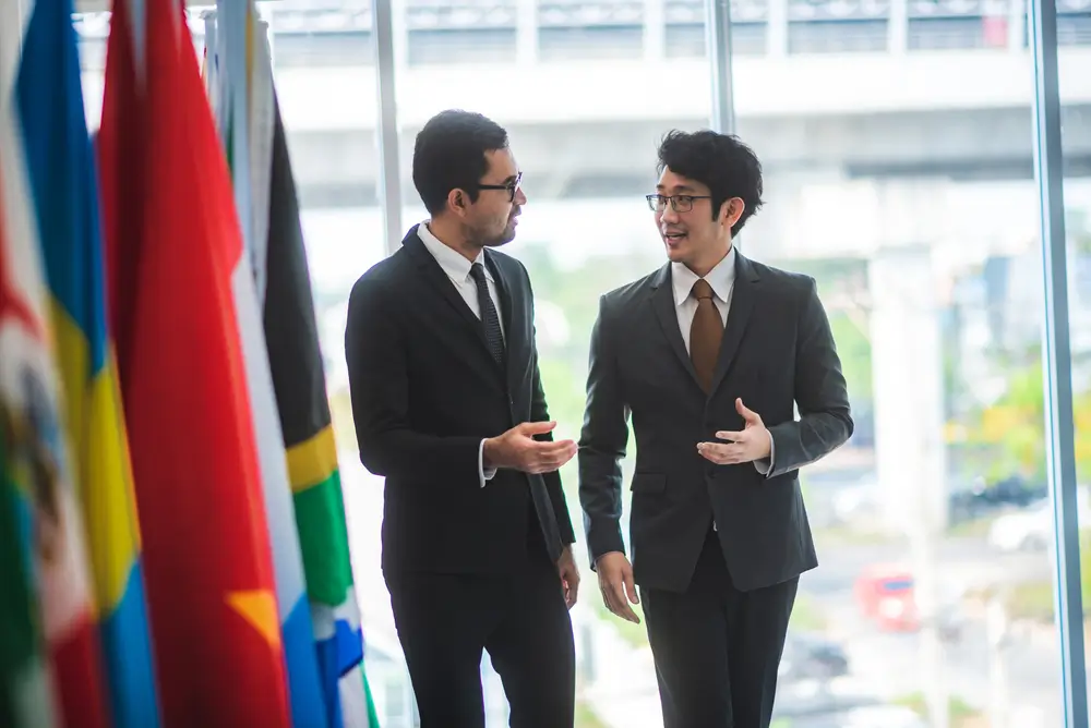 Two business professionals discussing international relations in a diplomatic setting with multiple national flags in the background.