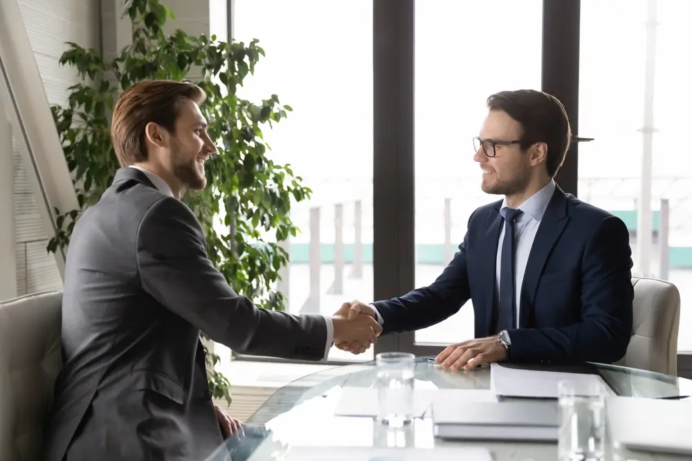 Two businessmen shaking hands after finalizing a government contracting deal in a professional office setting.