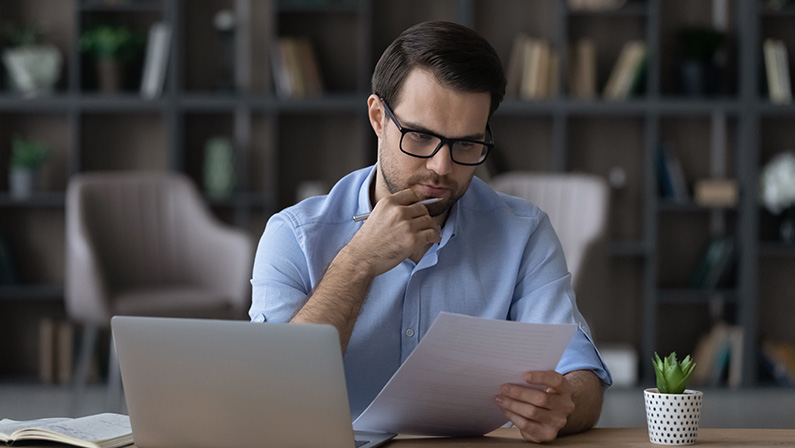 Working with papers. Serious millennial male entrepreneur work at home office alone hold read document hardcopy think on conditions terms. Focused young man engaged in paperwork edit text of agreement