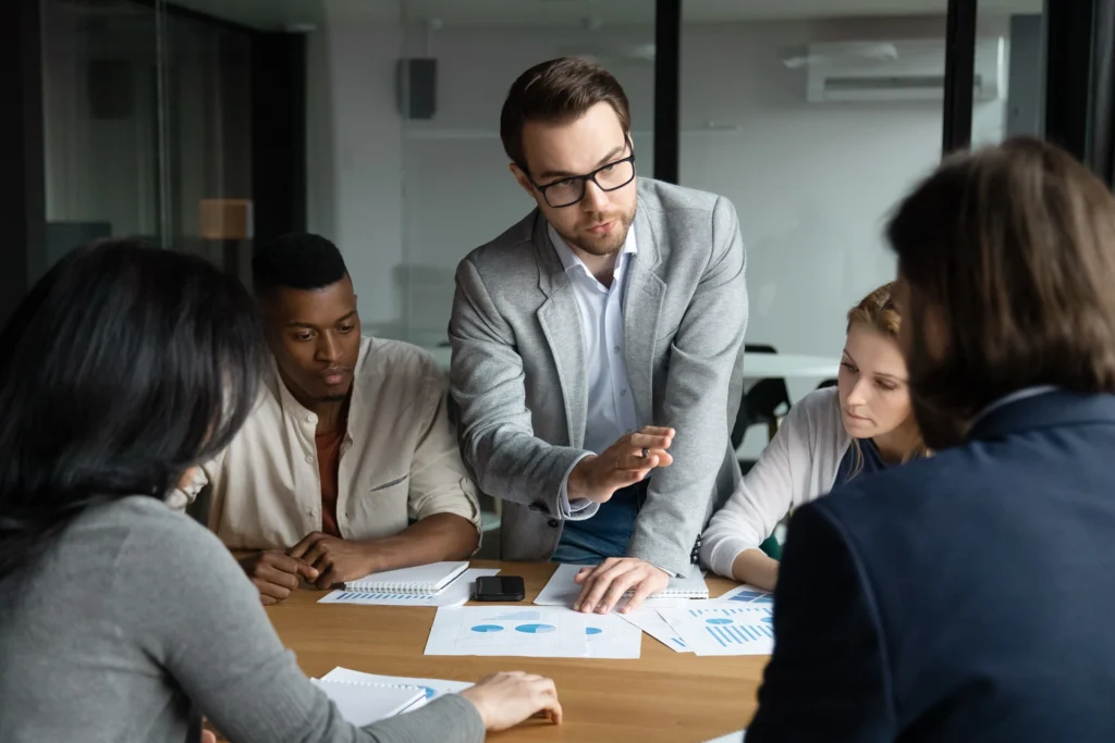 Focused confident young employee in eyeglasses giving arguments about marketing strategy or sharing own business ideas with focused motivated multiracial workgroup at brainstorming meeting in office.
