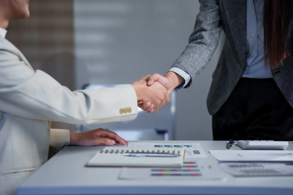 Two business professionals engage in a handshake across a desk, symbolizing agreement and partnership. Documents and graphs on the table highlight a professional environment